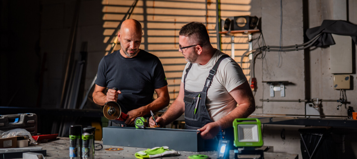 Produktový specialista RECA radí kolegovi při práci v dílně s ruční svítilnou a dalším nářadím na stole.