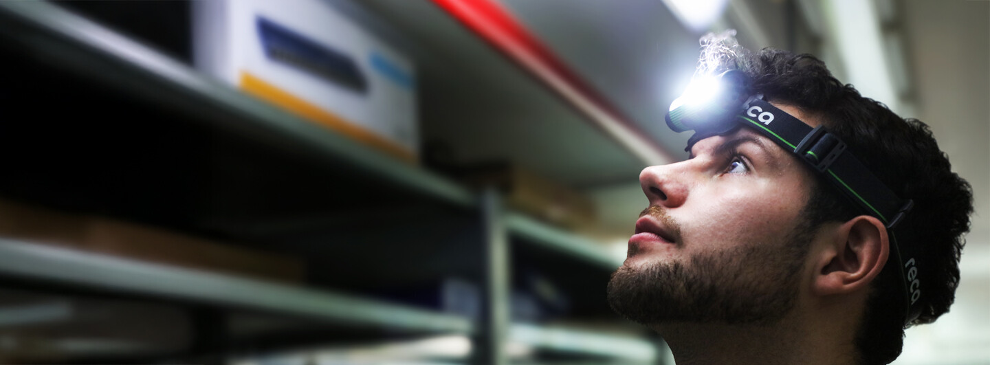 Pracovník ve skladu s nasazenou čelovkou RECA 5v1 při práci mezi regály.